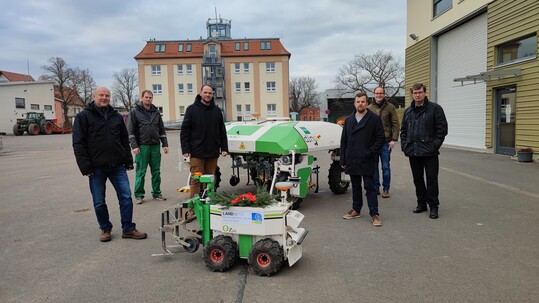 Coronakonforme und dennoch feierliche bergabe des Dino in Kllitsch, Sachsen: Den Schlssel zur Fern-Bedienung des Roboters berbrachte Dinos kleiner Bruder Oz (im Vordergrund). V. li. n. re.: Frank Nicklisch, BayWa Verkaufsberater Sachsen; Marvin Wenzel, Servicetechniker Smart Farming fr Sachsen; Cornelius Donath, Verkaufsleiter Europa Naio Technologies; Till Kunkel, Projektmitarbeiter Landnetz; Dr. Maximilian Deutsch, Leiter Stabstelle Digitalisierung beim LfULG; Dr. Uwe Bergfeld, Abteilungsleiter Landwirtschaft beim LfULG.  